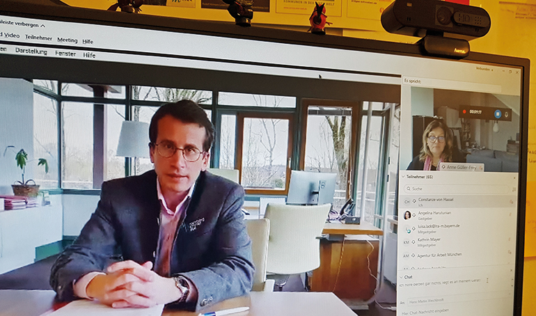 Landrat Stefan Frey sprach sein virtuelles Grußwort in seinem Amtszimmer; im Hintergrund Moderatorin Anne Güller-Frey von MigraNet. Bild: Bayerische GemeindeZeitung