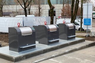 Unterirdisches Entsorgungssystem mit Betonbehälter im Erdreich: Ein Müllcontainer wird per Kranfahrzeug zur Entleerung angehoben, automatische Sicherheitsplattform verhindert Unfälle.